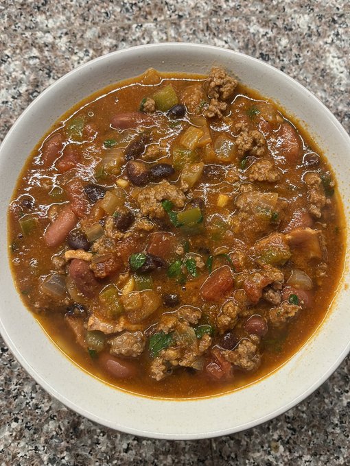 A top-down view of a bowl of hearty chili containing ground meat, beans, tomatoes, and green peppers, served in a white bowl on a granite countertop.