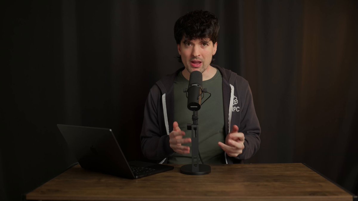 The image features a man sitting at a desk with a laptop computer, microphone, and speaker. The man appears to be giving a speech or presentation using the microphone and speaker setup. There are no other people visible in the image, but the focus is on the man's interaction with his equipment.