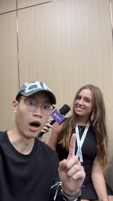 A selfie-style photo of two people at what appears to be a conference; an Asian man wearing glasses and a baseball cap makes an excited expression while pointing upward, standing next to a smiling woman in a black top who is holding a microphone with an "Agora" logo.