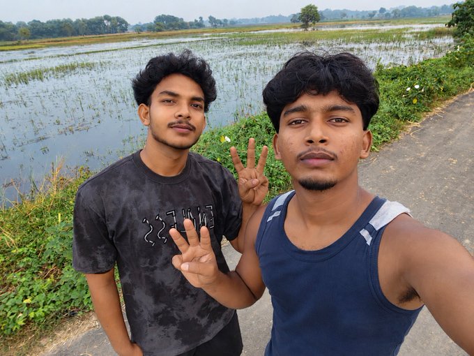 A selfie of two young men standing by a rural road next to a flooded rice paddy, with one making a peace sign.