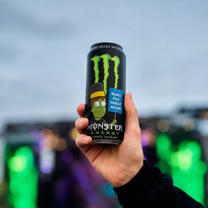 A hand holds up a black Monster Energy Zero Sugar can with a special design featuring a cartoon character in a top hat holding a sign that reads "MAKE FIAT GREAT AGAIN," set against a blurred concert or festival background.
