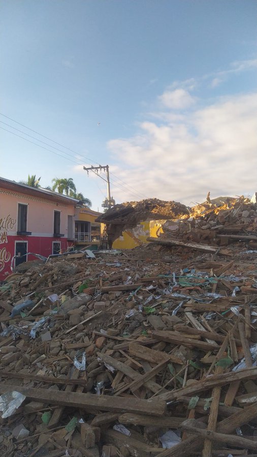 A photograph of a large pile of rubble and debris from a collapsed building in a town, with a pink building still standing on the left under a partly cloudy sky.