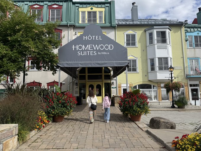 The entrance to Hôtel Homewood Suites by Hilton, featuring a black awning with the hotel's name, located in a village-like setting with colorful buildings and potted plants.