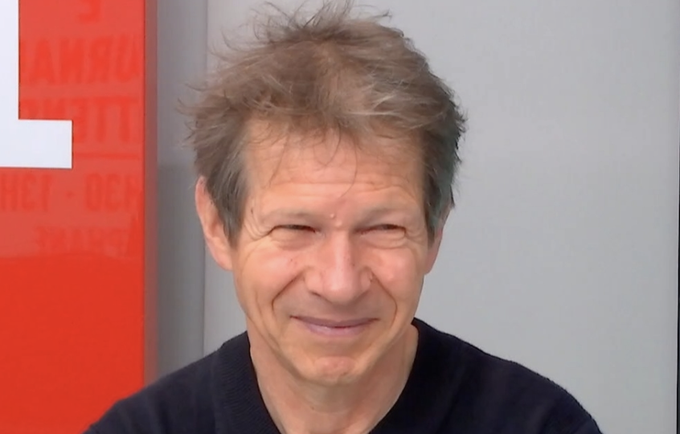 The image features a man with short hair wearing a black shirt, smiling for the camera. The background includes a red sign that reads "Lloyd's" and "Jancovici", which could possibly be related to the man or his work.