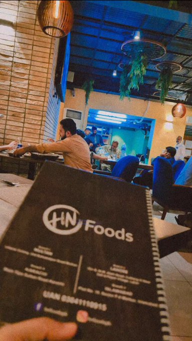 A first-person view of someone holding an 'HN Foods' menu at a restaurant with blue ambient lighting, hanging plants, and other diners visible in the background.