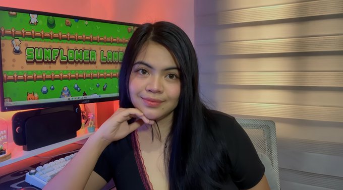 A woman with long dark hair sits in front of a computer monitor displaying a game called "SUNFLOWER LAND," posing for a photo in a room with red and blue ambient lighting.