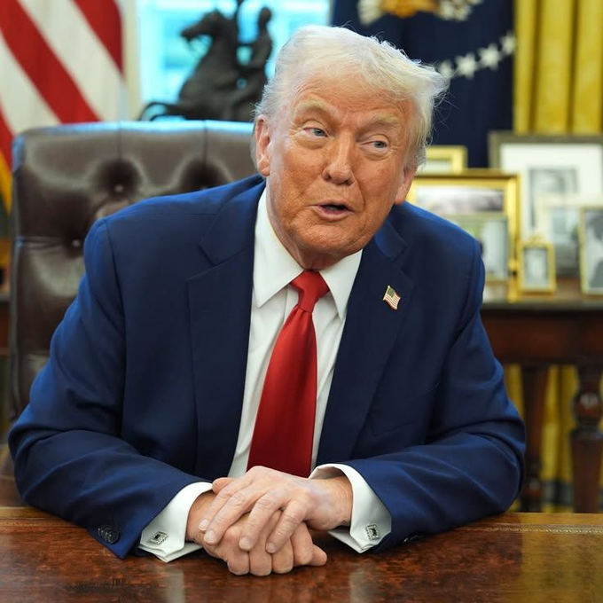 Donald Trump seated at the Resolute Desk in the Oval Office, wearing a navy blue suit and red tie, with the American flag visible behind him.