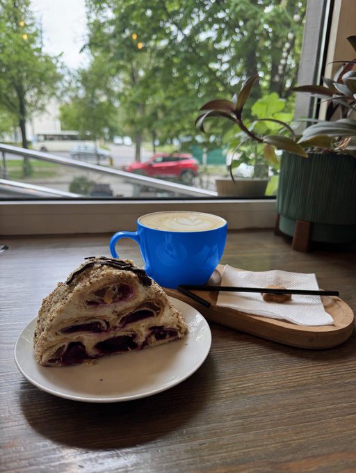 The image features a plate with a slice of cake, likely a pastry or dessert, placed on top of a wooden table. Next to the plate, there's a cup filled with coffee or tea, possibly a cappuccino or latte. The background shows a window overlooking trees and cars passing by outside. The post that accompanies this image reads "Gm for the base culture".