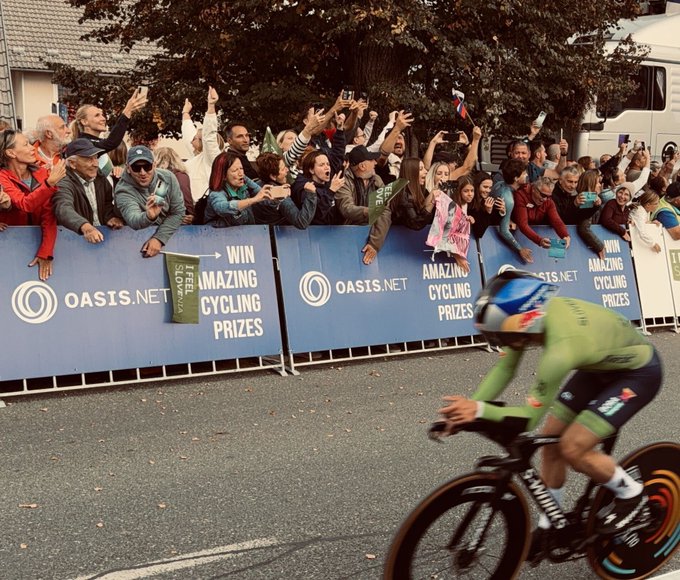 A cyclist in green racing gear speeds past cheering fans behind barriers advertising 'OASIS.NET' and 'WIN AMAZING CYCLING PRIZES'.