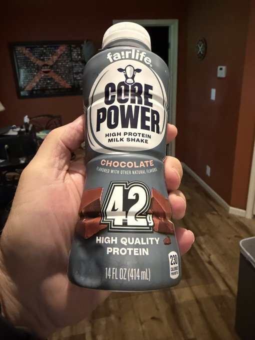 A hand holds up a bottle of Fairlife Core Power High Protein Milk Shake in chocolate flavor, featuring 42g of high-quality protein and 250 calories per 14 fl oz (414 mL) bottle, fitting the post's fitness-focused message about protein consumption.