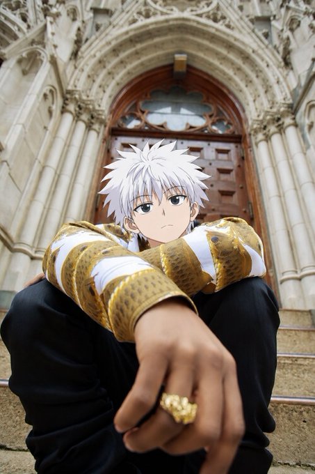 A cosplayer styled as the anime character Killua Zoldyck with white spiky hair and a gold-patterned jacket, sitting on steps before a Gothic cathedral doorway.