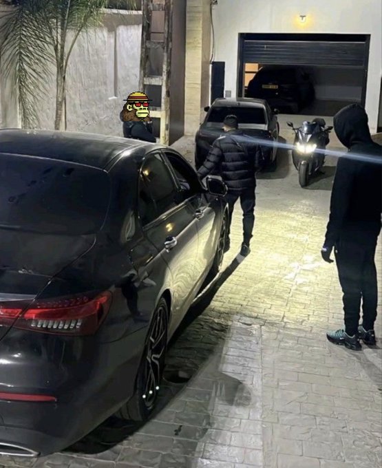 A nighttime outdoor scene showing a black luxury sedan parked on a paved driveway near an open garage. Several figures in dark clothing are visible, with one person's face obscured by a cartoon burger sticker.