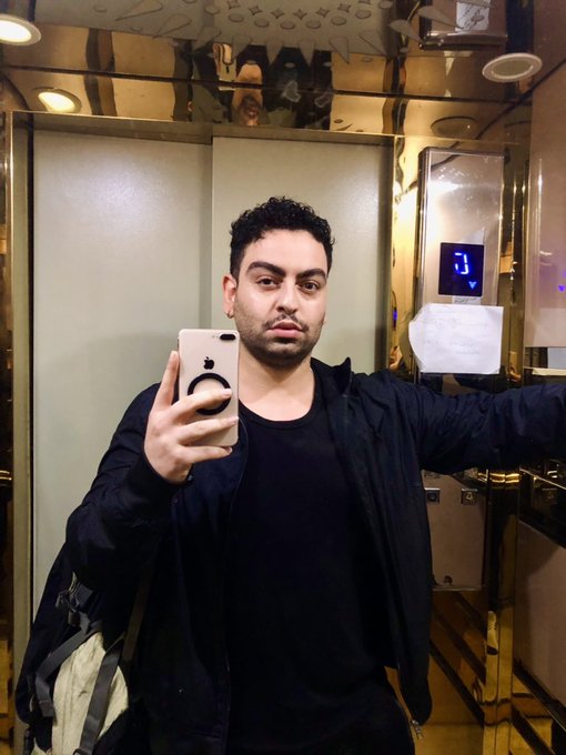 A man with dark curly hair taking a mirror selfie in an ornate elevator with gold-colored interior panels, wearing a black shirt and dark jacket.