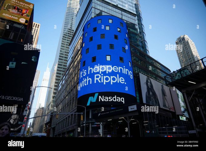 A large blue Nasdaq digital billboard in an urban setting displays the text 'It's happening with Ripple' along with the Nasdaq logo.