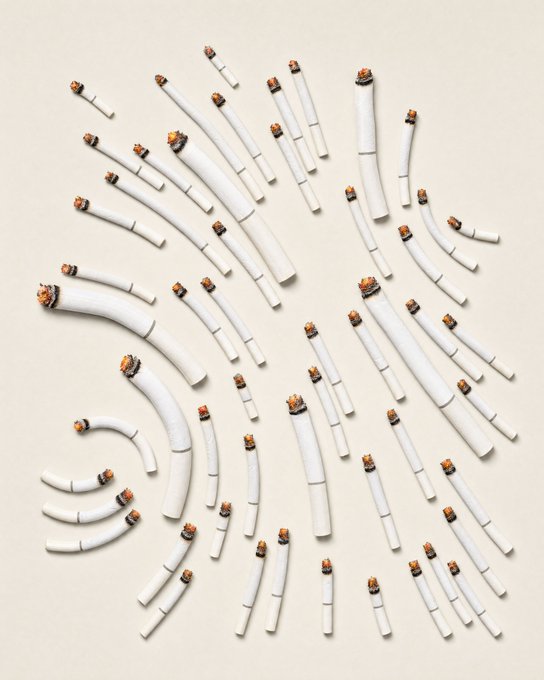 An overhead artistic photograph shows numerous lit cigarettes arranged in a scattered pattern on a plain light background, each broken into pieces.