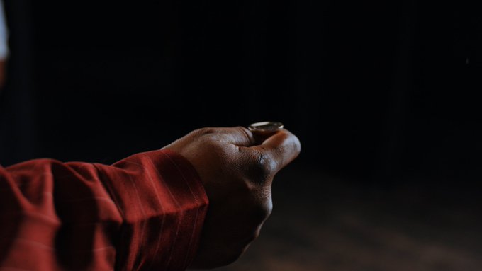 A hand wearing a red sleeve extends a single coin against a dark, shadowed background, capturing a quiet moment of hope or fortune. The image visually echoes the caption's message, suggesting how something small—a single coin—can hold the power to transform one's destiny.