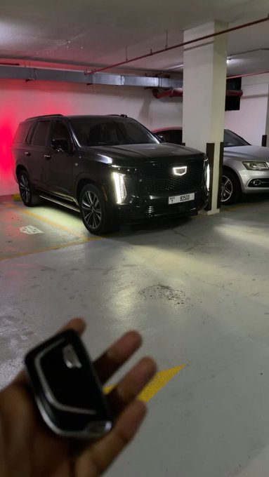A black Cadillac Escalade parked in a parking garage with its headlights on, as a hand holds a key fob in the foreground.