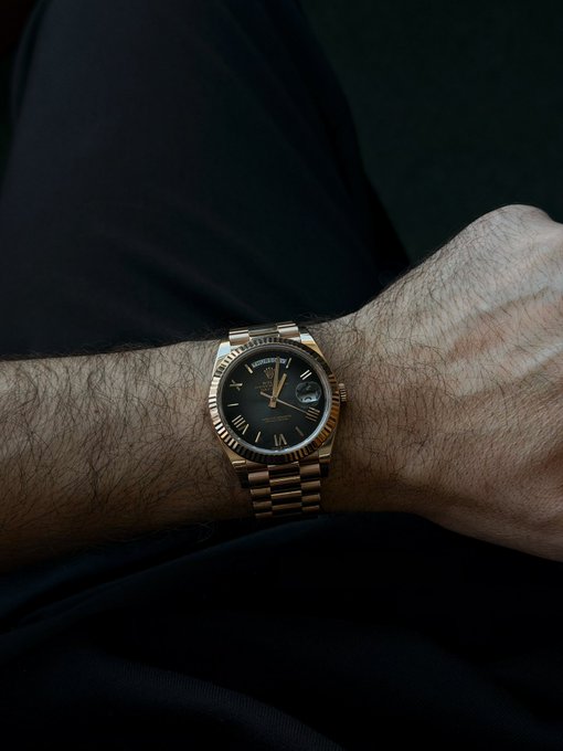 A close-up of a hairy wrist wearing a gold Rolex Day-Date watch with a black dial and Roman numerals against dark fabric.