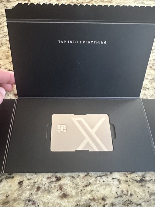 A sleek black metal card in an open black presentation box, featuring an embossed 'X' logo and chip, with the tagline 'TAP INTO EVERYTHING' printed inside the lid.