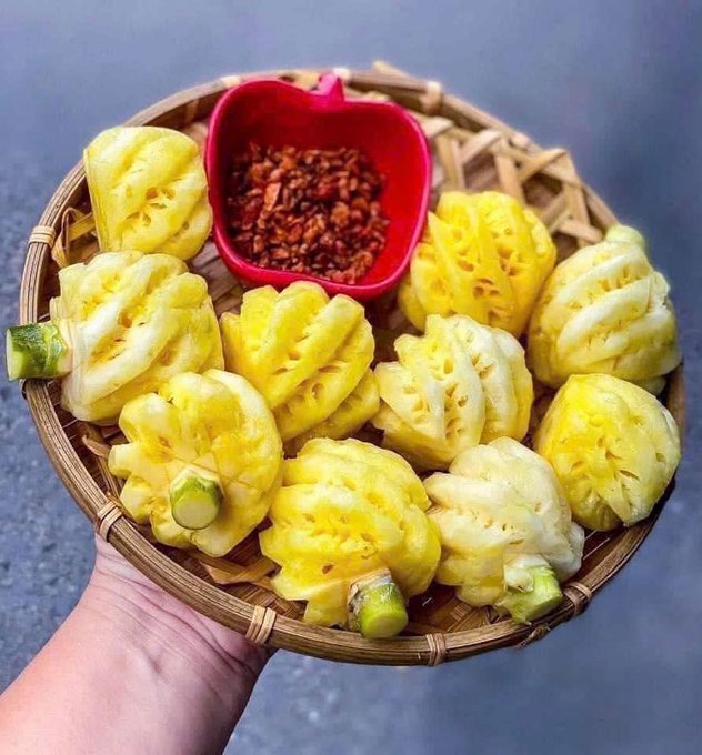 A hand holding a woven basket filled with carved yellow pineapple pieces, accompanied by a small red dish of red chili flakes.