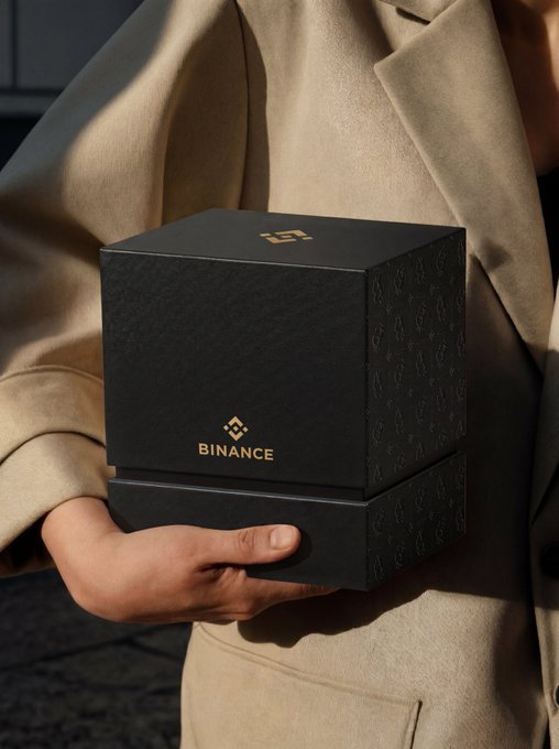 A person in a beige coat holds a black gift box branded with the "BINANCE" logo and cryptocurrency-themed patterns, teasing the contents of the mystery box in celebration of Binance's 9th anniversary.