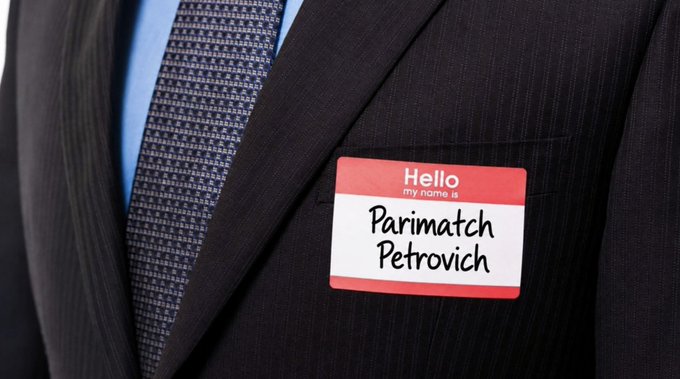 Close-up of a person wearing a dark pinstripe suit and blue patterned tie, with a red and white 'Hello my name is' sticker reading 'Parimatch Petrovich' on their lapel.