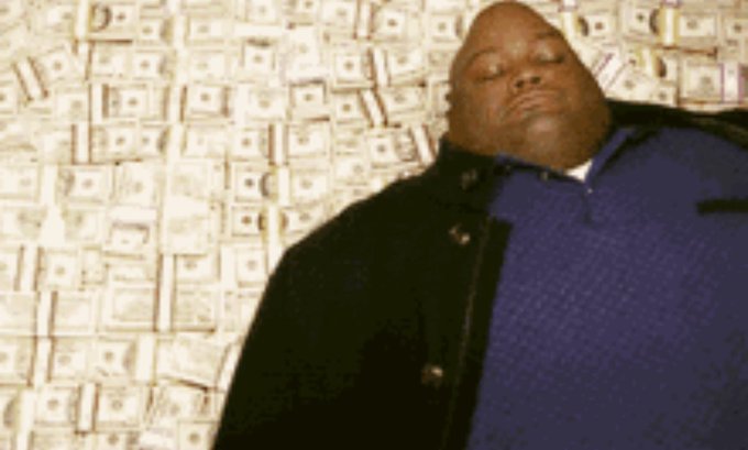 A man in a blue shirt and black jacket stands in front of a wall covered with stacks of money.