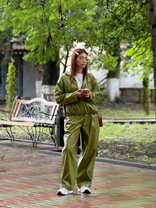 A young woman with red hair and glasses stands on a wet brick pathway in a park, wearing a matching olive-green tracksuit and holding her phone, appearing relaxed as she checks messages—perhaps scrolling through crypto tips on Jupiter Academy (@jup_academy) to plan her weekend of gym sessions, baking, and learning.