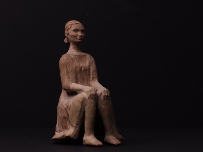 A small, rustic clay figurine of a seated woman with her hands resting on her knees, photographed against a solid black background.