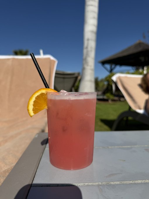 A pink cocktail garnished with an orange slice and black straw sits on a patio table under a sunny blue sky, with lounge chairs and a palm tree in the background.
