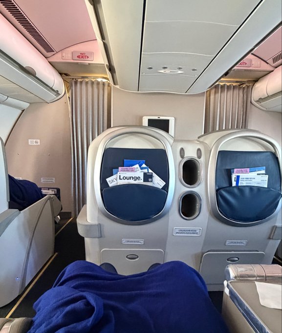 Interior view of an airplane cabin looking forward at the back of business or first-class seats with "Lounge" cards in the pockets, featuring cream and grey upholstery, and a passenger's blue blanket-covered legs in the foreground.