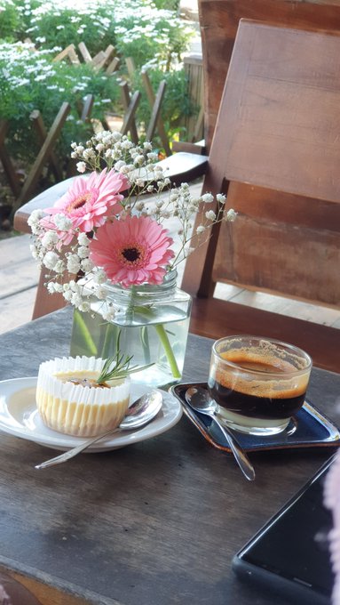 A wooden table holds a glass jar with pink gerbera daisies and baby's breath, a small cupcake on a white plate, and an espresso with cream beside a spoon. The serene setting of flowers, sweets, and coffee perfectly matches the calm, relaxing Sunday morning vibe described in the Vietnamese caption about recharging for the week ahead.