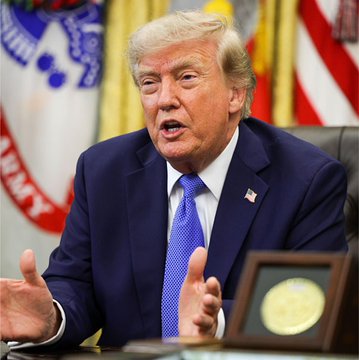 Donald Trump sits at a desk, gesturing with both hands as he speaks, wearing a navy blue suit and a blue tie with an American flag pin. In the background, a flag with the word "ARMY" visible on a red ribbon is seen to the left.