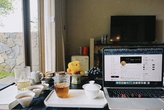 A cozy workspace setup with a laptop displaying a Chinese-language webpage, alongside tea cups, a teapot, and a small yellow character figurine on the table.