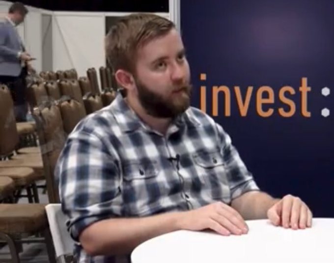 A bearded man in a blue and white plaid shirt sits at a table being interviewed, with the word "invest" visible on a purple banner behind him, illustrating a discussion about Schwab's offering of crypto assets including $BTC, $ETH, $AVAX, $SOL, and $LINK.