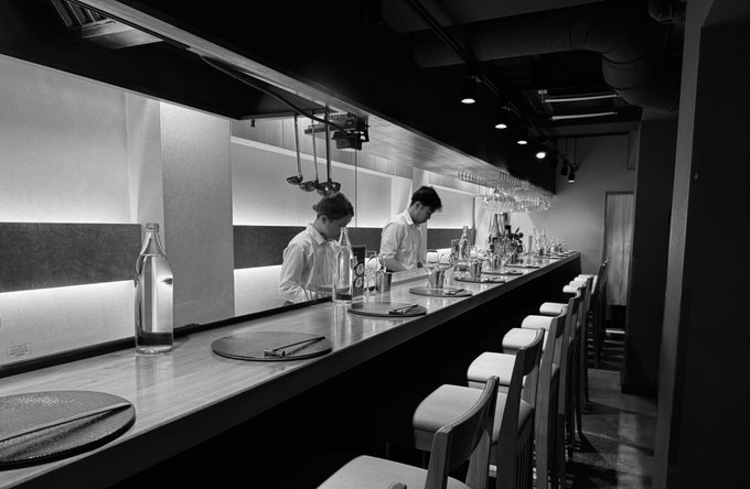 A black and white photograph of two chefs working behind a counter at a minimalist sushi restaurant, with bar stools lined up along the counter and glassware hanging overhead.