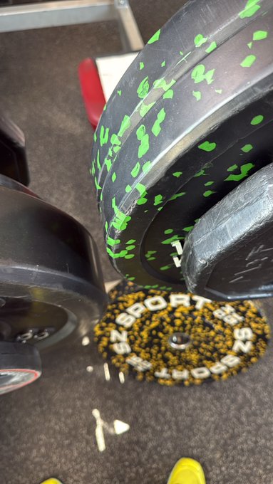 The image shows a pair of legs wearing yellow shoes standing next to two black weightlifting machines with green and white designs, one of which has a green sticker on it. The text "gQuack" appears in the image as well.