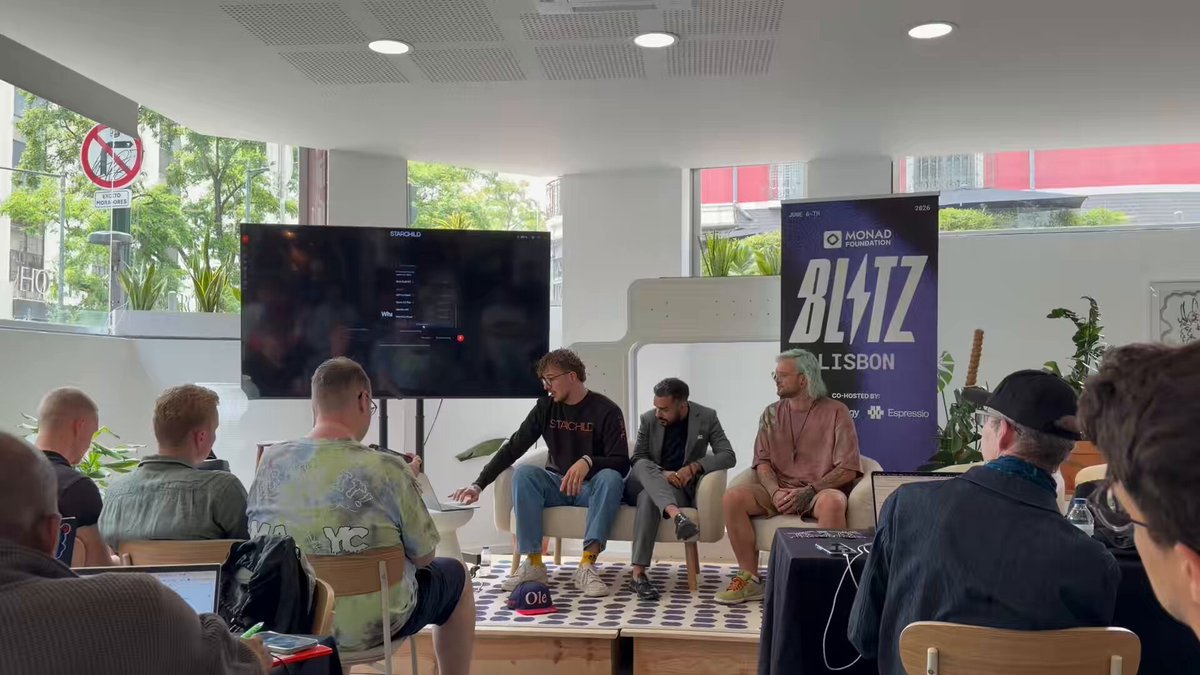 The image shows a group of people sitting around a table, with some men wearing black jackets and others wearing green shirts. They are engaged in conversation or listening to a presentation. The room has a large TV screen mounted on the wall behind them, which is displaying information related to Starchild, an AI platform. There is also a banner hanging from the ceiling that reads "Urban".