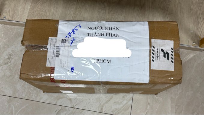 A cardboard shipping box with a partially redacted label addressed to "THANH PHAN" in "PHCM," bearing a QR code and a barcode sticker, sits on a tiled floor.