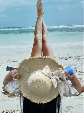 A person lies on a sunny beach wearing a straw hat with a bow, their legs raised in the air, holding a phone and a water bottle, with the watermark '@Dreame' visible.