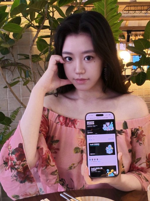 A young woman in a pink floral off-shoulder top holds up a smartphone displaying a prediction market app while posing in a plant-filled café setting.

The image connects to the post by visually showcasing a user engaging with a mobile prediction market platform, illustrating the accessibility of BitMart's featured prediction market that allows low-entry participation starting at just 2 USDT.