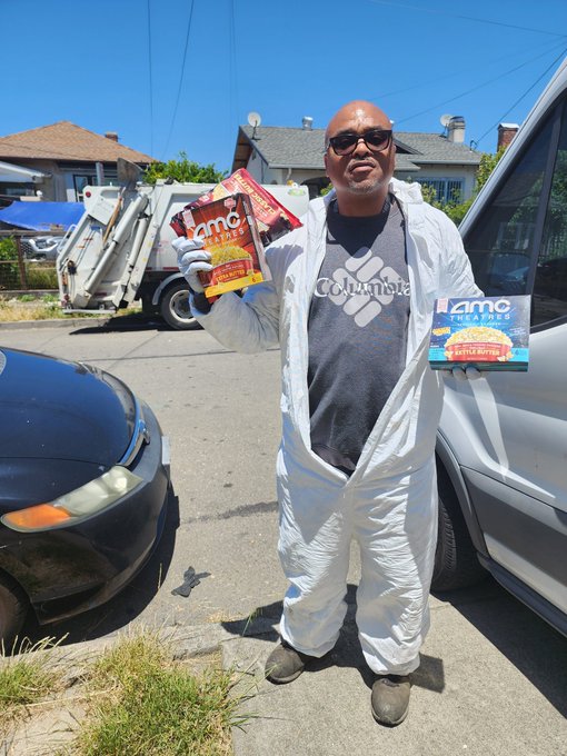 iced coffee shop owner, dressed in white and black, holds two boxes of coffee on a sidewalk with a car parked nearby.