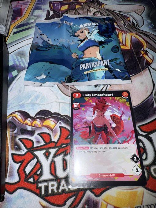 The image features a promotional item for an anime-themed event, with two items displayed side by side - one being a card and the other a book cover featuring a character from the same anime series. The text on the card reads "Particpants", while the book cover displays a character named "Lady Emburhart".