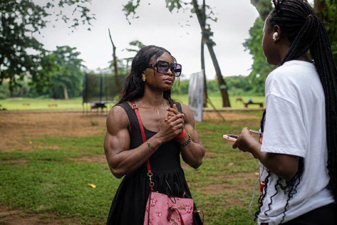 A muscular woman with long dark hair, wearing sunglasses, a black top, and a floral pink bag, stands on a grassy area talking to another person. The second person has long braided hair, wears a white t-shirt and earbuds, and holds a phone.