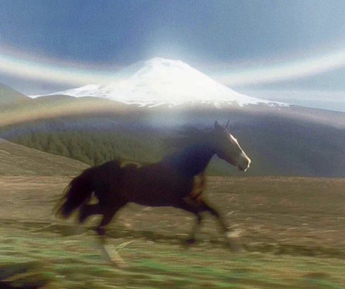 A blurry photo of a black horse galloping across a grassy field with a snow-capped mountain and rainbow in the background.