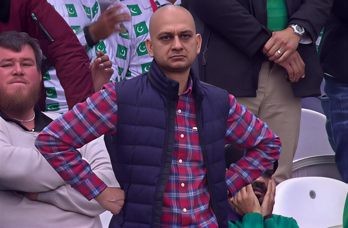 A bald man in a plaid shirt and blue vest stands with hands on hips among a crowd at what appears to be a sporting event.