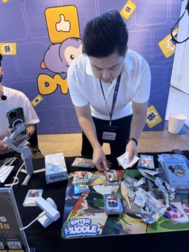 A man in a white shirt stands at a booth displaying merchandise including t-shirts, stickers, and toys labeled 'Enter the Huddle' with a cartoon character backdrop.