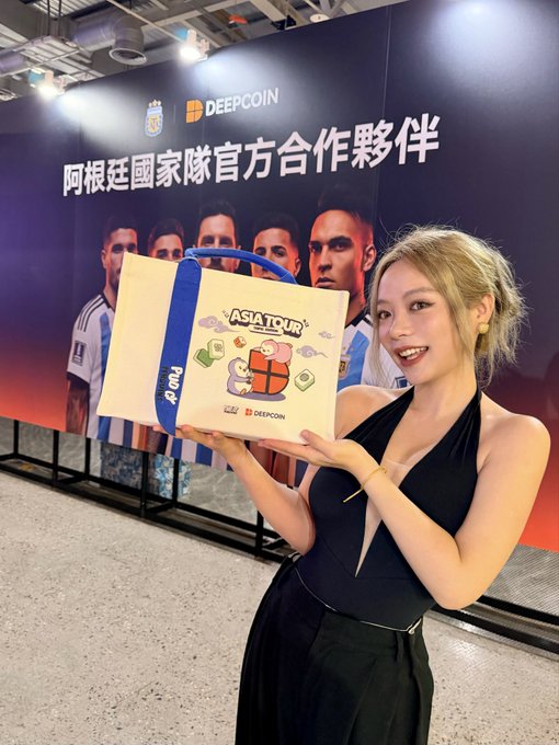 A woman in a black halter dress holds up a 'ASIA TOUR' gift box branded with 'DEEPCOIN' at an Argentina National Team event, standing in front of a backdrop reading '阿根廷国家隊官方合作夥伴' and 'DEEPCOIN'.