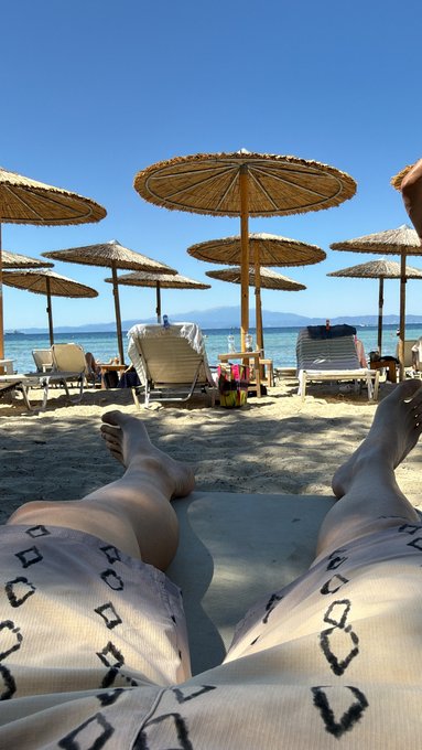A first-person perspective shot shows legs stretched out on a beach towel under a clear blue sky, with straw umbrellas, white sun loungers, the calm sea, and distant mountains in the background.