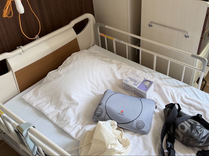 A hospital bed with white linens holds a silver Sony portable CD player, a box of tissues, a cream-colored bag, and a black backpack on the mattress.